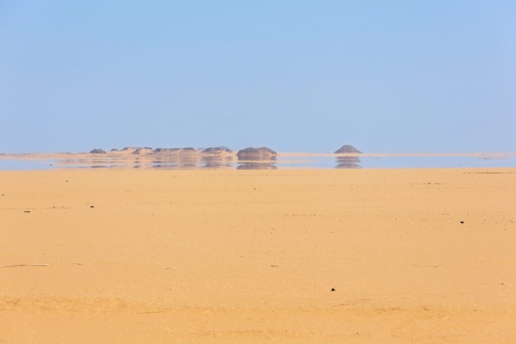 Expansive sandy desert landscape with distant rocky outcrops and clear blue sky
