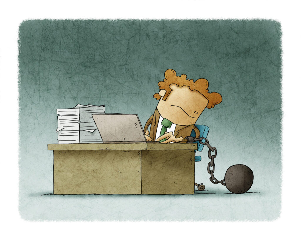 Cartoon illustration of a tired office worker chained by the leg to a ball while sitting at a desk with a laptop and piles of papers